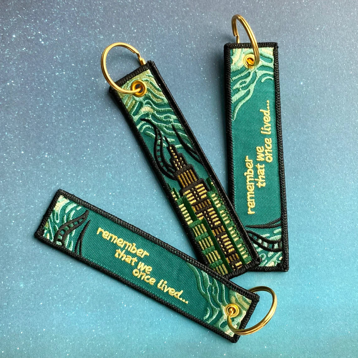 Two green keychains with gold text and Amaurot cityscape pattern on a blue background