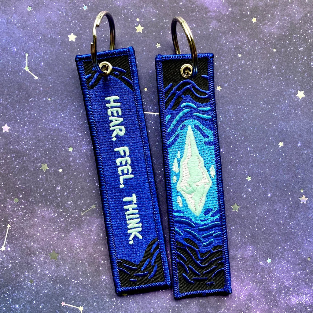 Two blue embroidered keychains with mothercrystal design and HEAR. FEEL. THINK. on a starry background